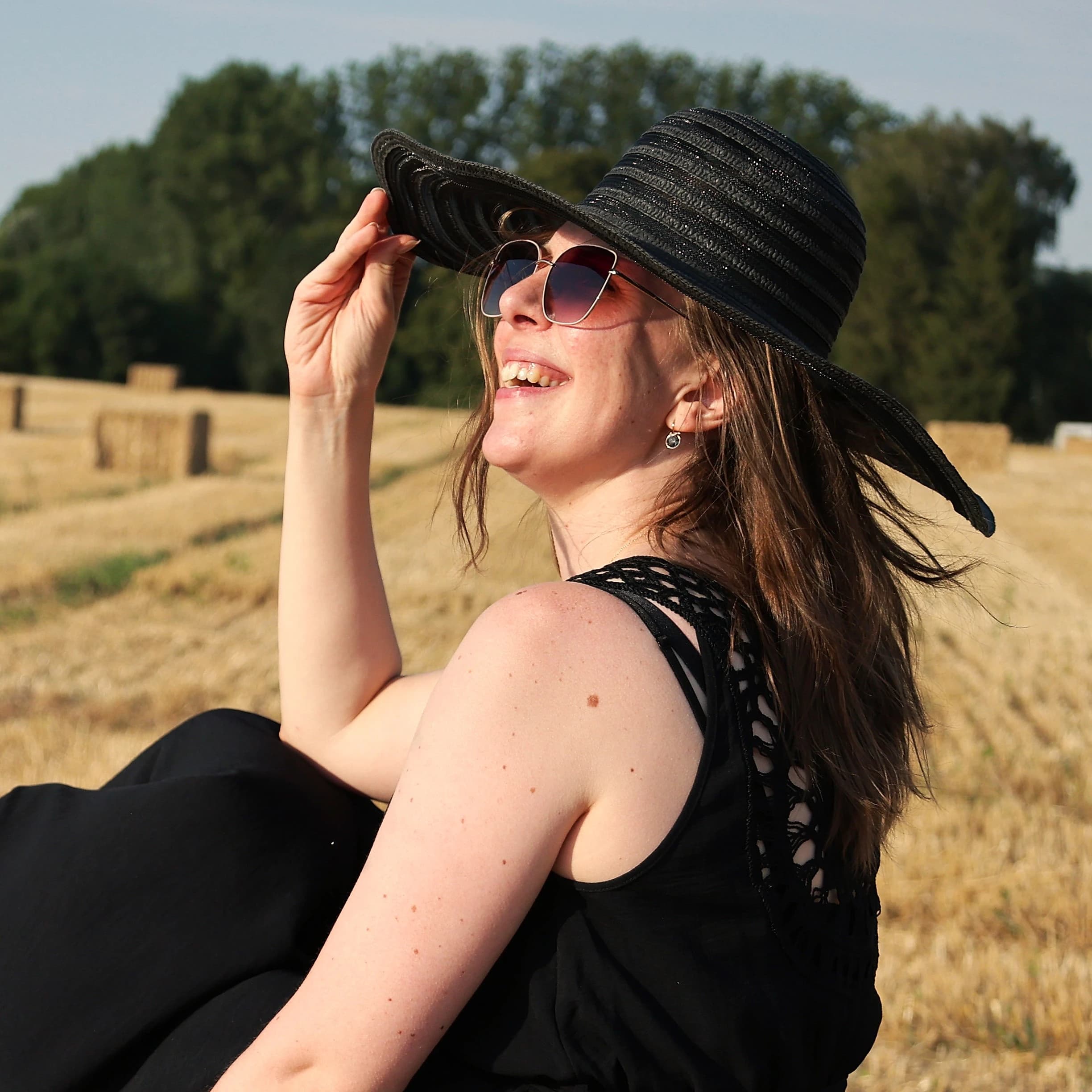Femme souriante portant un chapeau noir élégant dans un champ de blé ensoleillé.